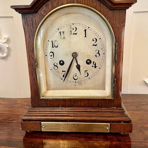 Puede incluir: Un reloj de sobremesa de madera vintage con una esfera color crema y números negros. El reloj tiene un bisel color latón y un marco de madera marrón oscuro. La esfera del reloj marca las 5:27. Una pequeña placa de latón está en la base.