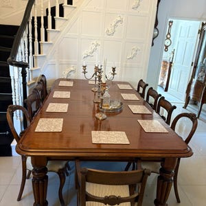 May include: A grand, polished wooden dining table with eight chairs. The table has a dark brown finish and is set with placemats, a candelabra, and glassware. The chairs are a mix of styles, some with curved backs and others with striped seats.