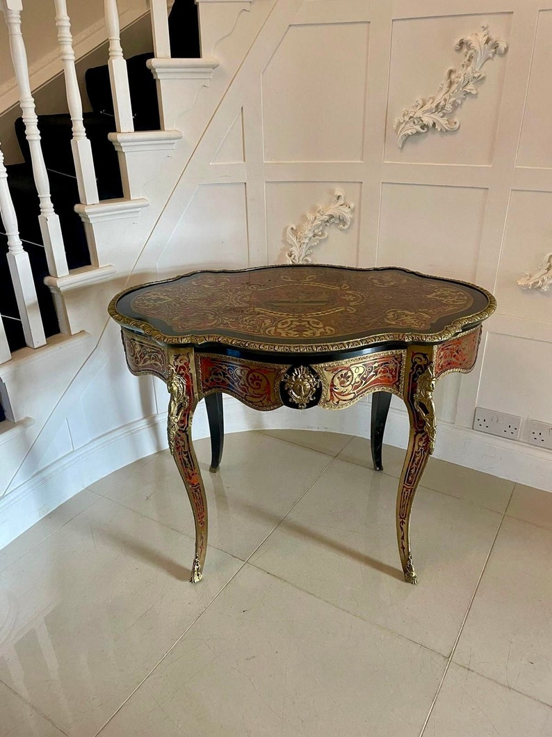 Puede incluir: Una mesa de madera negra e incrustada en oro con un caj&oacute;n y patas ornamentadas. La mesa tiene un borde festoneado y una tapa de marqueter&iacute;a.