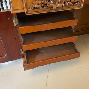 May include: A wooden cabinet with a carved drawer and four open shelves. The cabinet is made of light wood and has a dark wood finish. The shelves are empty.
