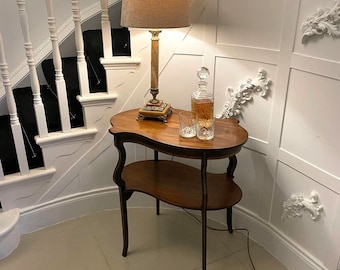 May include: A brown wooden side table with two shelves and a curved top. A lamp with a beige lampshade and a glass decanter with a stopper are on the table.
