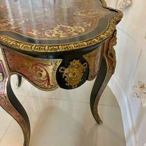 Puede incluir: Una mesa auxiliar antigua de color negro y dorado con intrincados detalles de marqueter&iacute;a y un dise&ntilde;o floral tallado. La mesa tiene una parte superior curva y tres patas.