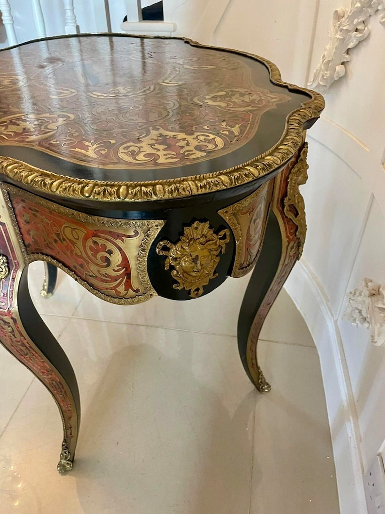 Puede incluir: Una mesa auxiliar antigua de color negro y dorado con intrincados detalles de marqueter&iacute;a y un dise&ntilde;o floral tallado. La mesa tiene una parte superior curva y tres patas.