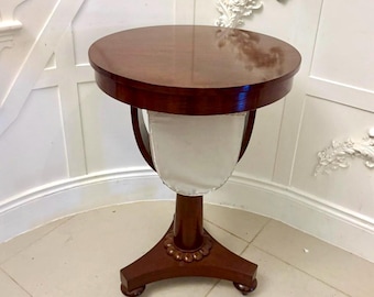 May include: A small, round, mahogany side table with a glossy finish. The table has a pedestal base with a decorative floral design and three feet. A white fabric panel is suspended beneath the tabletop.
