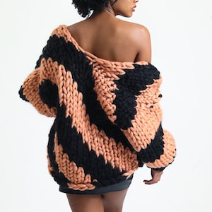 May include: A black and peach striped chunky knit cardigan with a relaxed fit. The cardigan is draped over one shoulder and the model is wearing a black mini skirt.