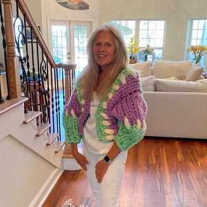 Könnte beinhalten: Eine Frau trägt ein weißes Hemd und eine weiße Jeans mit einem grob gestrickten Cardigan in Lila- und Grüntönen. Sie steht auf einem gemusterten Teppich in einem häuslichen Umfeld.