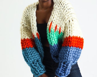 May include: A chunky knit cardigan with a white base and colorful stripes in red, orange, green, and blue. The cardigan has a relaxed fit and a slightly cropped length.
