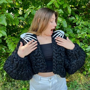 K&ouml;nnte beinhalten: Ein schwarzer, kurzer Cardigan mit grobmaschiger Strickstruktur und wei&szlig;en Skelett-Hand-Applikationen an den Schultern.