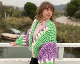May include: A vibrant, chunky knit cardigan in green, lilac, and white. The oversized sweater features a unique stitch pattern. The sleeves and body are a mix of colours, creating a bold and cosy look. The cardigan is a statement piece.