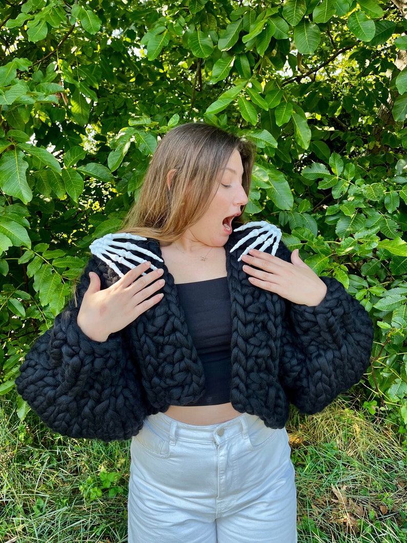 K&ouml;nnte beinhalten: Ein schwarzer, kurzer Cardigan mit grobmaschiger Strickstruktur und wei&szlig;en Skelett-Hand-Applikationen an den Schultern.