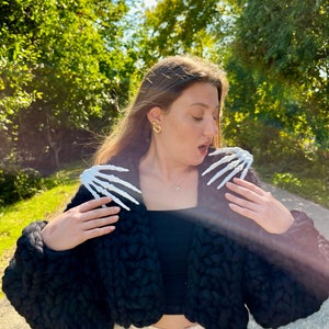 K&ouml;nnte beinhalten: Eine Frau tr&auml;gt einen schwarzen, grob gestrickten Cardigan mit wei&szlig;en Skeletth&auml;nden auf den Schultern. Sie tr&auml;gt eine wei&szlig;e Hose und hat ihr Haar offen.