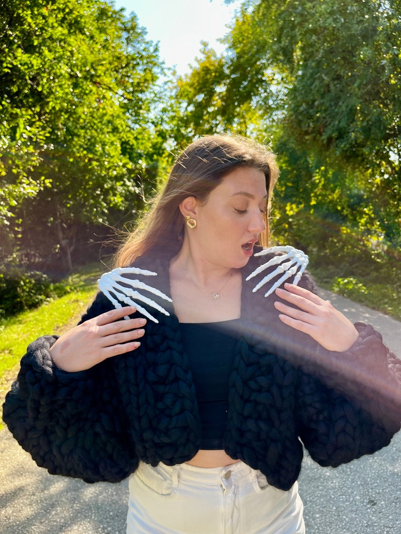 K&ouml;nnte beinhalten: Eine Frau tr&auml;gt einen schwarzen, grob gestrickten Cardigan mit wei&szlig;en Skeletth&auml;nden auf den Schultern. Sie tr&auml;gt eine wei&szlig;e Hose und hat ihr Haar offen.