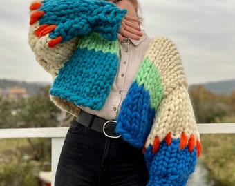 May include: A chunky knit cardigan with a colorful striped pattern in shades of blue, green, white, and orange. The cardigan has a cropped length and long sleeves.