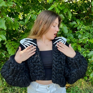 K&ouml;nnte beinhalten: Ein schwarzer, grob gestrickter Cardigan mit wei&szlig;en Skeletth&auml;nden an den Schultern. Der Cardigan ist kurz geschnitten und hat eine lockere Passform.
