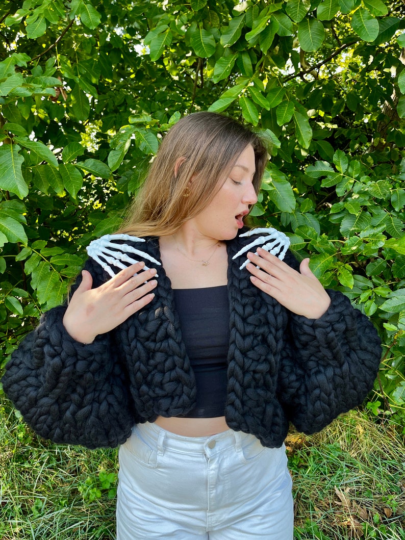 K&ouml;nnte beinhalten: Ein schwarzer, grob gestrickter Cardigan mit wei&szlig;en Skeletth&auml;nden an den Schultern. Der Cardigan ist kurz geschnitten und hat eine lockere Passform.