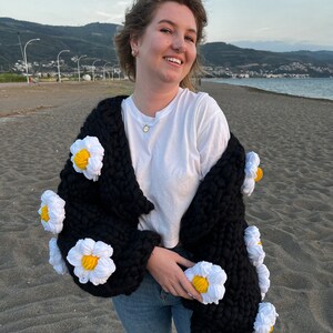 K&ouml;nnte beinhalten: Ein schwarzer Strickcardigan mit wei&szlig;en, glockenf&ouml;rmigen Verzierungen an den &Auml;rmeln. Der Cardigan wird &uuml;ber einem wei&szlig;en T-Shirt und einer blauen Jeans getragen.