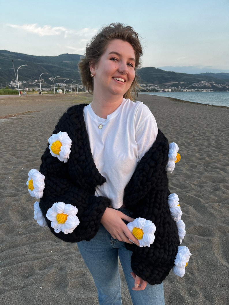 K&ouml;nnte beinhalten: Ein schwarzer Strickcardigan mit wei&szlig;en, glockenf&ouml;rmigen Verzierungen an den &Auml;rmeln. Der Cardigan wird &uuml;ber einem wei&szlig;en T-Shirt und einer blauen Jeans getragen.
