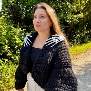 K&ouml;nnte beinhalten: Ein schwarzer, grob gestrickter Cardigan mit wei&szlig;en Skeletth&auml;nden auf den Schultern. Der Cardigan ist kurz geschnitten und hat eine lockere Passform.