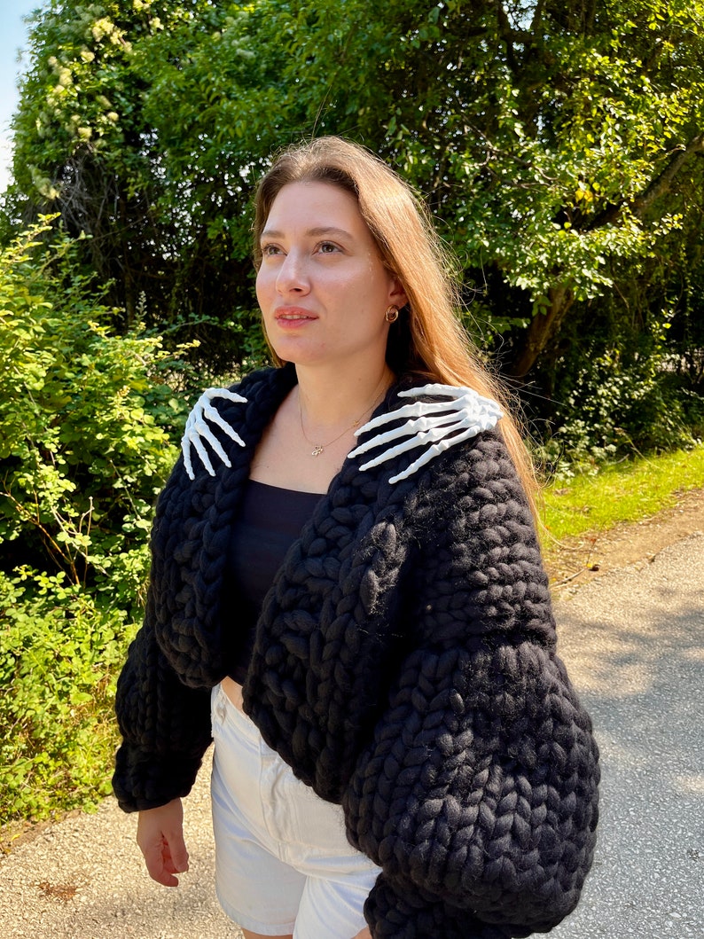 K&ouml;nnte beinhalten: Ein schwarzer, grob gestrickter Cardigan mit wei&szlig;en Skeletth&auml;nden auf den Schultern. Der Cardigan ist kurz geschnitten und hat eine lockere Passform.