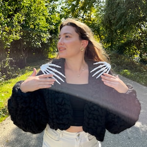 K&ouml;nnte beinhalten: Ein schwarzer, kurzer Cardigan mit grobmaschiger Strickstruktur. Der Cardigan hat wei&szlig;e Skeletth&auml;nde auf den Schultern. Die Person, die den Cardigan tr&auml;gt, steht auf einem gepflasterten Weg.