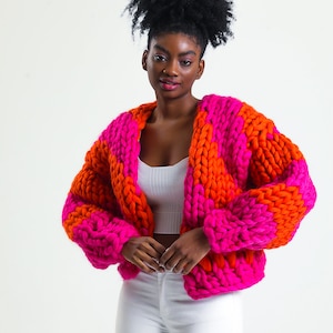 May include: A pink and orange chunky knit cardigan with a loose fit. The cardigan is made of thick yarn and has a relaxed, casual style.