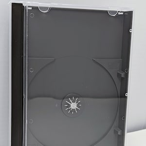 May include: A clear plastic CD case with a black spine and a black disc holder. The case is empty.