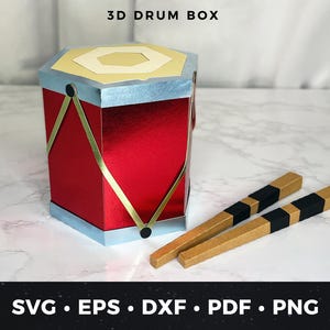 Op de afbeelding: Een rode 3D-drumbox met gouden en zilveren accenten en twee drumstokken. De doos heeft bovenaan de tekst "3D DRUM BOX". De onderkant van de afbeelding heeft de tekst "SVG • EPS • DXF • PDF • PNG".