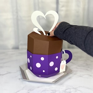 May include: A purple and brown papercraft teacup with white polka dots and a white heart-shaped steam design. The teacup has a white tag with a pink heart on it.