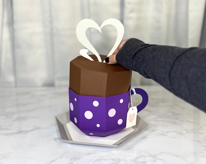 May include: A purple and brown papercraft teacup with white polka dots and a white heart-shaped steam design. The teacup has a white tag with a pink heart on it.