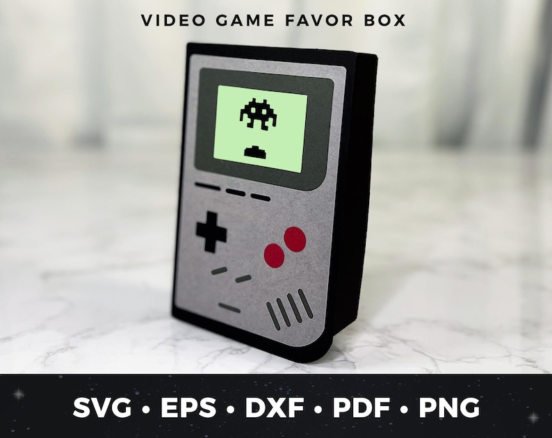 May include: A gray and black video game favor box shaped like a retro handheld game console. The box has a green screen with a pixelated alien spaceship on it. The box has red buttons and a plus sign button.