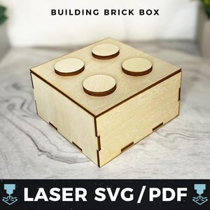 May include: A wooden box shaped like a building brick with four indented circles on the top. The box is made of light brown wood and has a natural wood finish.  BUILDING BRICK BOX is printed on the top of the image.