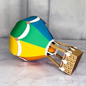 Puede incluir: Un globo aerost&aacute;tico de papel colorido con una cesta marr&oacute;n. El globo es una mezcla de amarillo, naranja, verde y azul. Se ven detalles blancos y dorados brillantes. La cesta tiene un patr&oacute;n decorativo.