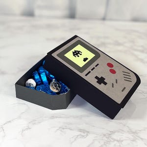May include: A black and gray cardboard box shaped like a vintage handheld video game console. The box is open and filled with blue and silver wrapped chocolate candies. The console has a yellow screen with a pixelated alien spaceship graphic.