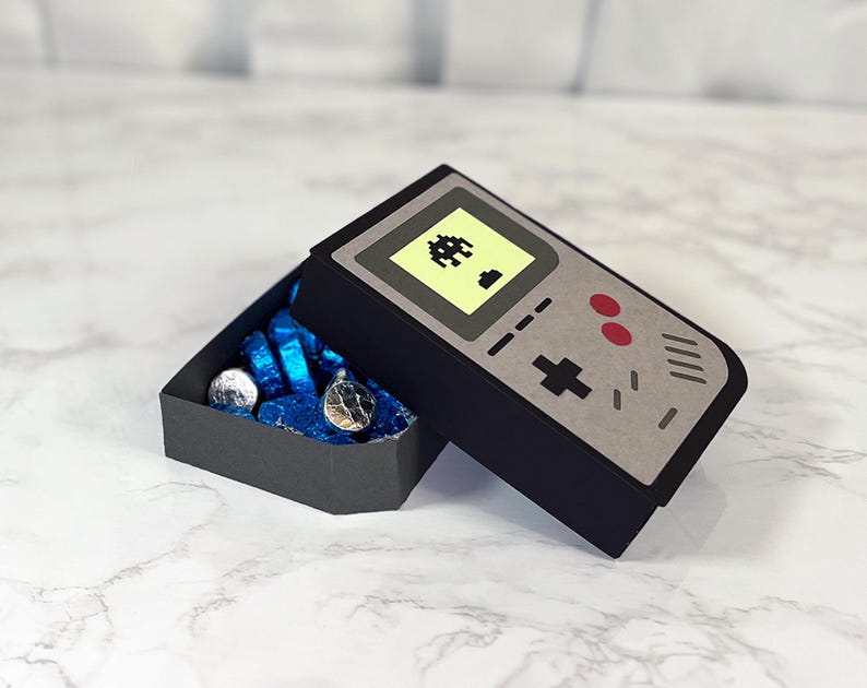 May include: A black and gray cardboard box shaped like a vintage handheld video game console. The box is open and filled with blue and silver wrapped chocolate candies. The console has a yellow screen with a pixelated alien spaceship graphic.
