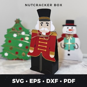 Puede incluir: Una caja de cascanueces roja y dorada con una cara blanca y un sombrero negro. La caja está decorada con botones dorados y un lazo de cuerda marrón. La caja está sobre una superficie blanca. El texto "NUTCRACKER BOX" está en la parte superior de la imagen.