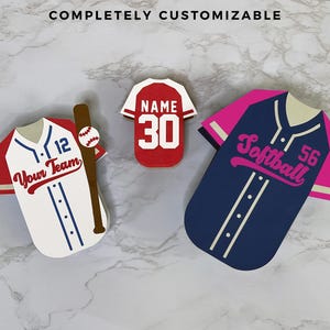 K&ouml;nnte beinhalten: Drei Baseball- und Softball-Dekorationen in Trikotform. Ein wei&szlig;es Trikot mit roten &Auml;rmeln und dem Text "Your Team" und einem Baseballschl&auml;ger. Ein rotes Trikot mit der Nummer 30. Ein marineblaues Trikot mit rosa &Auml;rmeln und dem Wort "Softball".
