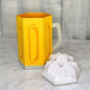 May include: A yellow paper beer mug with a white handle and a separate white paper foam element. The mug has a hexagonal shape with curved cutouts. The foam element is also hexagonal.