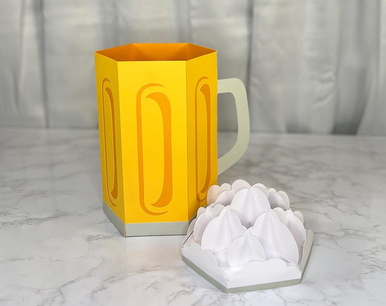 May include: A yellow paper beer mug with a white handle and a separate white paper foam element. The mug has a hexagonal shape with curved cutouts. The foam element is also hexagonal.