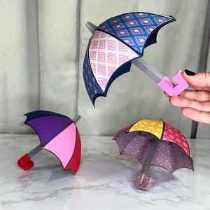 May include: Three paper umbrellas with different patterns and colors. The umbrellas are made of paper and have a pink, purple, and yellow and red color scheme. The umbrellas are on a white surface.
