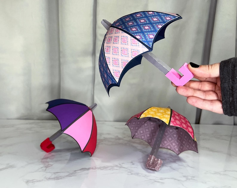 May include: Three paper umbrellas with different patterns and colors. The umbrellas are made of paper and have a pink, purple, and yellow and red color scheme. The umbrellas are on a white surface.