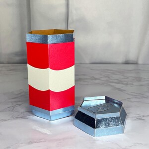 May include: A red and white striped cardboard tube with a silver top and bottom. The tube is open at the top and has a silver lid on the side. The lid is hexagonal in shape.