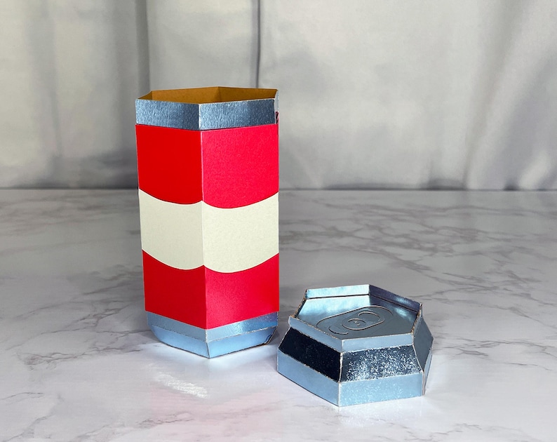 May include: A red and white striped cardboard tube with a silver top and bottom. The tube is open at the top and has a silver lid on the side. The lid is hexagonal in shape.
