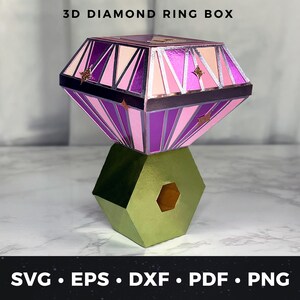 May include: A 3D diamond ring box with a geometric design. The top of the box is a diamond shape with pink, purple, and gold accents. The base is a green hexagon shape. The text "3D DIAMOND RING BOX" is at the top.