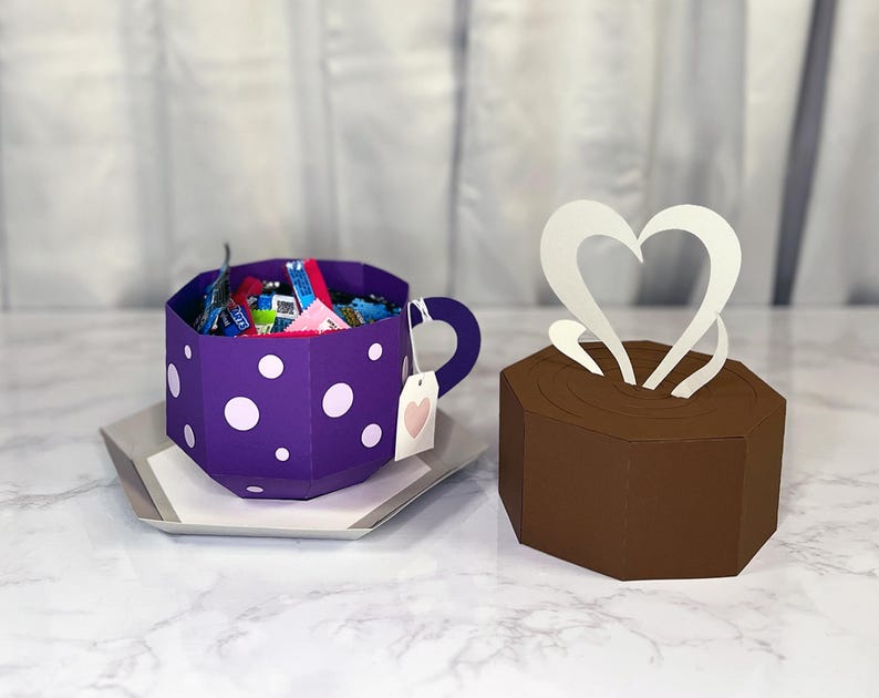 May include: A purple teacup with white polka dots filled with colorful candy. The teacup has a handle and a pink heart tag. A brown box with a white heart shaped steam design sits next to the teacup.