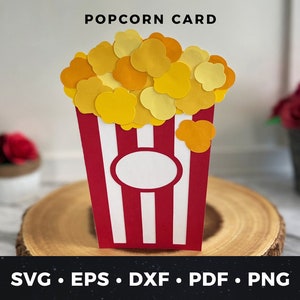 May include: A red and white striped popcorn box with yellow popcorn pieces on top. The box has a white circle in the centre. The text "POPCORN CARD" is at the top of the image. The image is on a wooden surface.