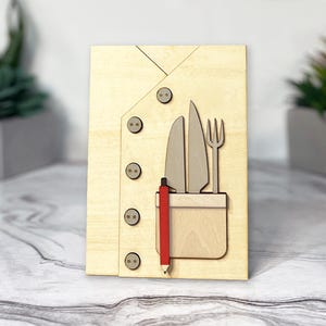 May include: A wooden cutout of a chef's jacket with a pocket containing a knife, fork, and pencil. The jacket has four buttons.