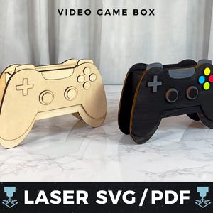 May include: Two wooden video game controller boxes are shown, one in natural wood and the other black. The controllers feature button and directional pad details. The text "VIDEO GAME BOX" and "LASER SVG/PDF" are visible.