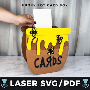 May include: A brown card box shaped like a honey pot with yellow honey dripping down the sides. Black text on the box reads "CARDS". The box has a yellow rim and is decorated with black bees. The text "HUNNY POT CARD BOX" is at the top.