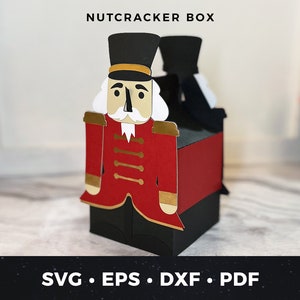 May include: A red and black nutcracker box with a white beard and gold trim. The box is made of paper and is designed to be assembled. The box is decorated with a nutcracker design and the words "NUTCRACKER BOX" are printed on the top.