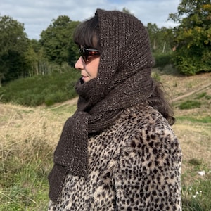May include: A dark brown knitted hood scarf with a textured, speckled yarn finish, worn over a faux fur coat featuring a black and tan leopard print pattern. The hood covers the head and drapes down into a long scarf section around the neck.